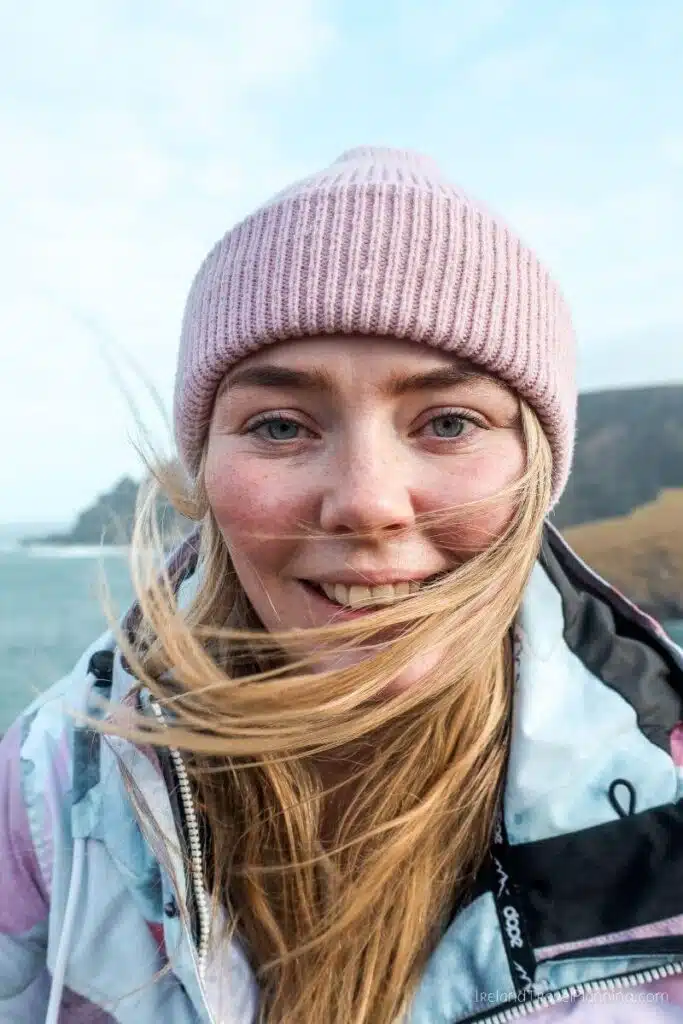 Young woman with blonde hair and blue eyes wearing a pink beanie and jacket, smiling outdoors.