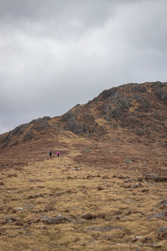 Two hikers ascend a grassy mountain slope under a cloudy sky.