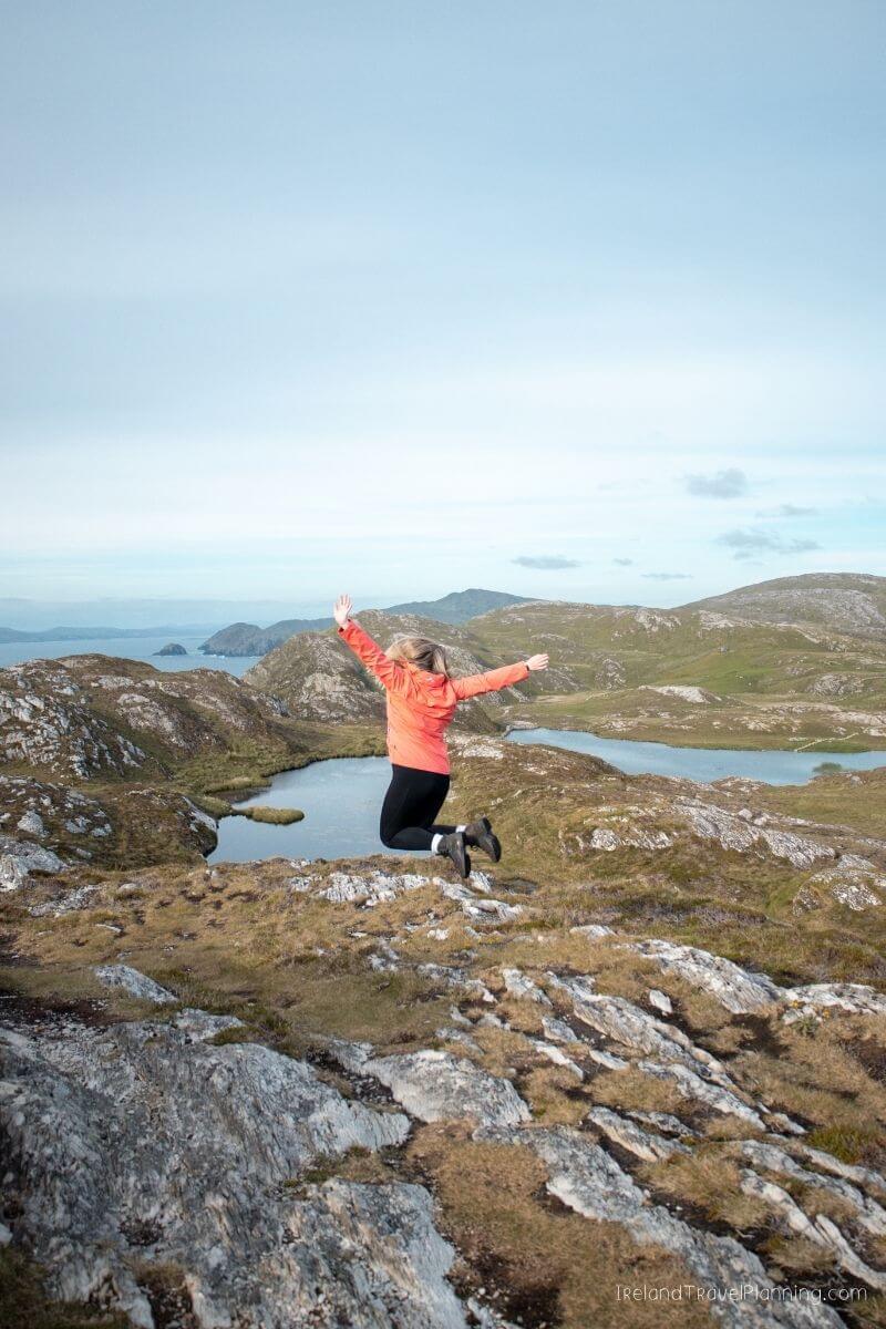 Woman in orange jacket jumping on rocky hill overlooking a lake and mountains