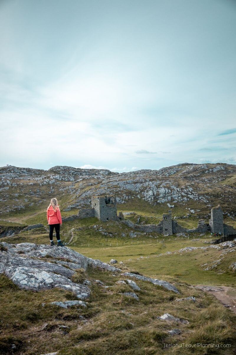 Woman in orange jacket overlooks ancient stone ruins in a rugged Irish landscape.