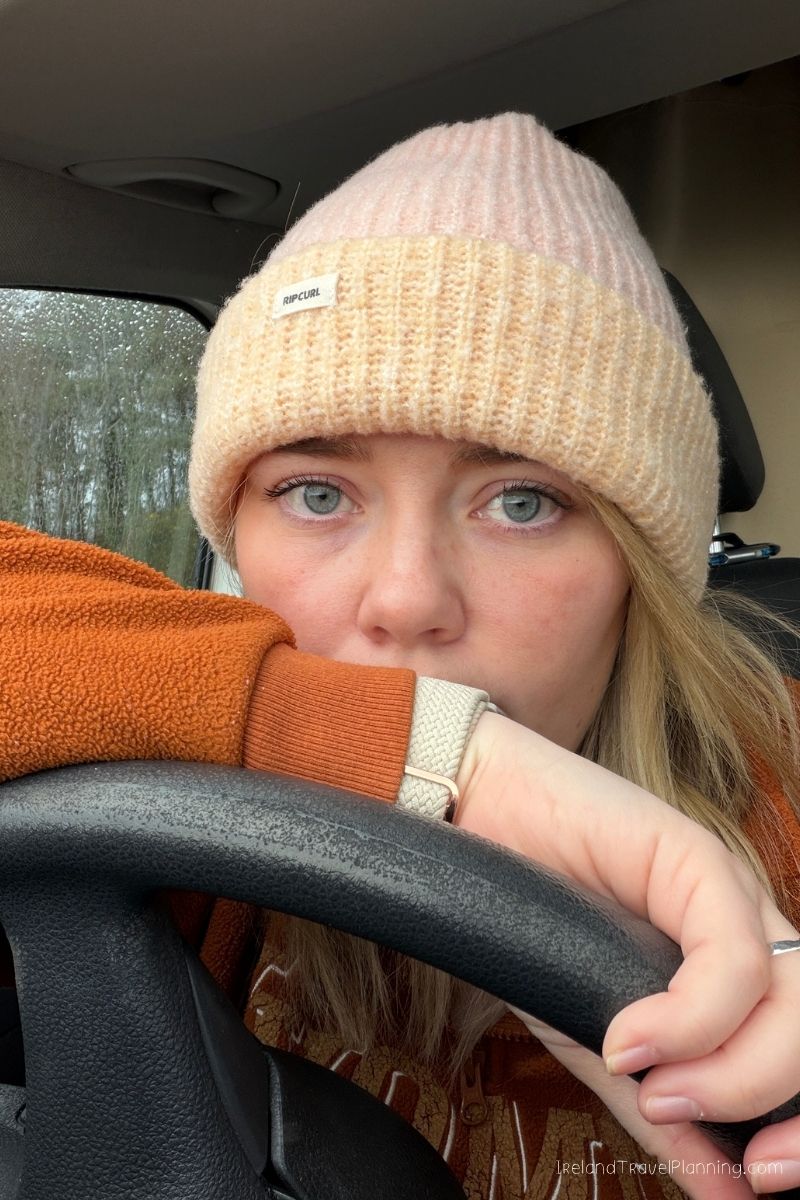 Woman in beanie and orange jacket in car, considering renting a car in Ireland.