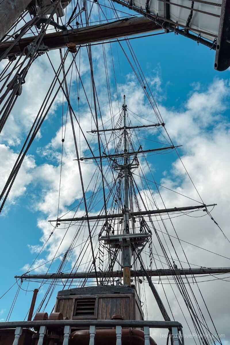View from below of a tall ship's mast and rigging against a blue sky with clouds.