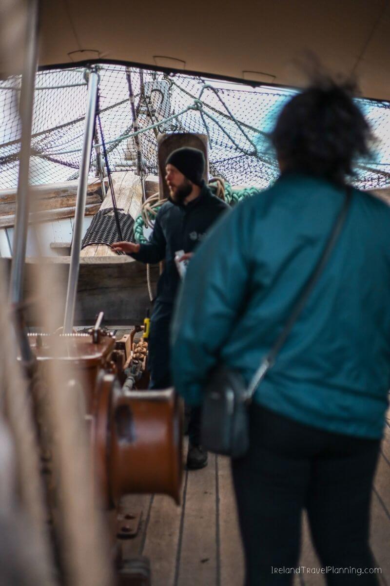 Man in beanie on a boat explains something to a person in a teal jacket
