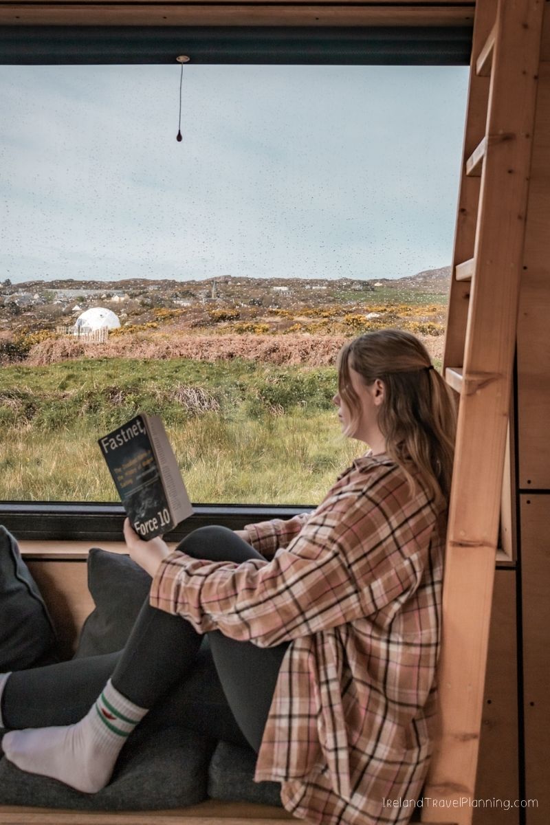 Woman reading "Fastnet Force 10" in a cozy cabin with a view of the Irish landscape