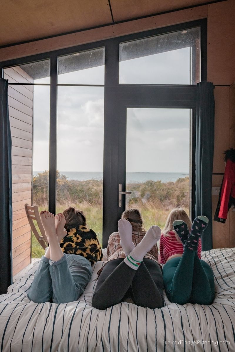 Three people relax on a bed, looking out a large window at the Irish coast.