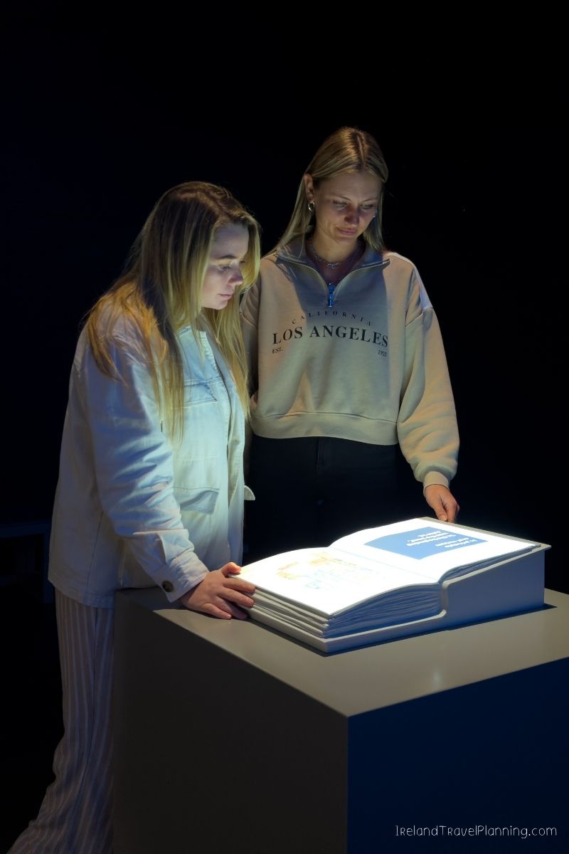 Two women examine a large open book at an exhibit, possibly related to an Ireland trip cost.
