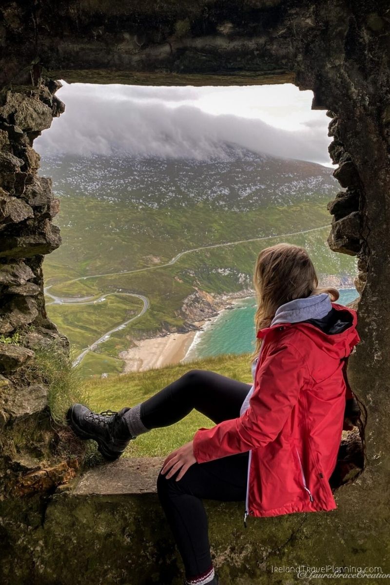 Woman in red jacket looks out from stone window at Irish coast and mountains.