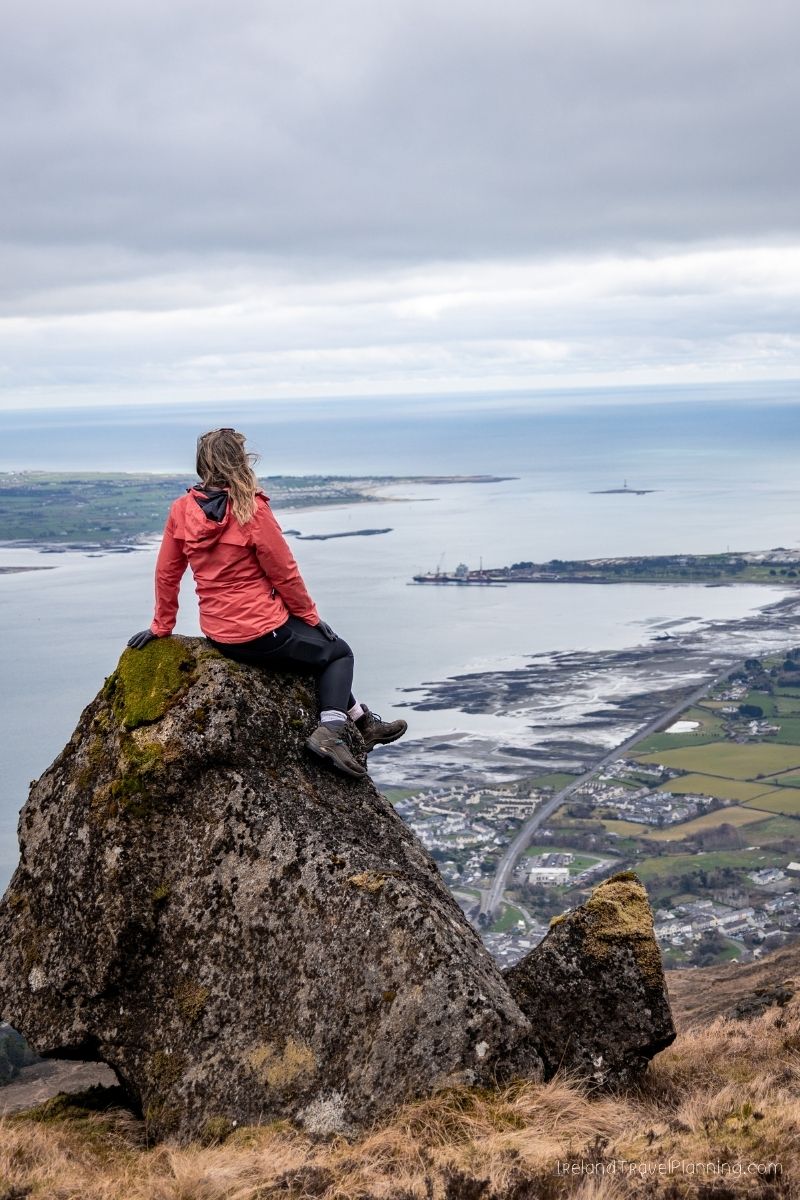 Woman in red jacket sits on mossy rock overlooking coastal town and sea