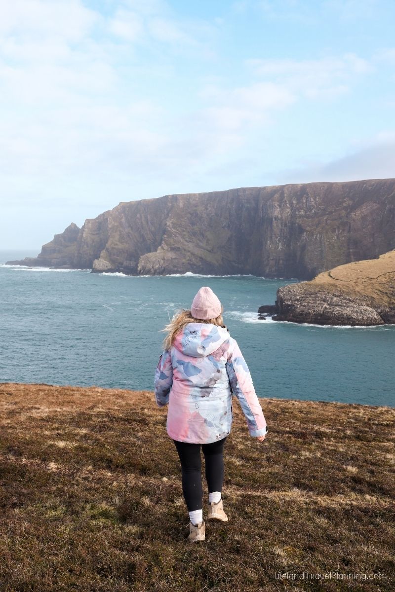 Woman in a pink hat and floral jacket looking at dramatic cliffs and ocean in Ireland.