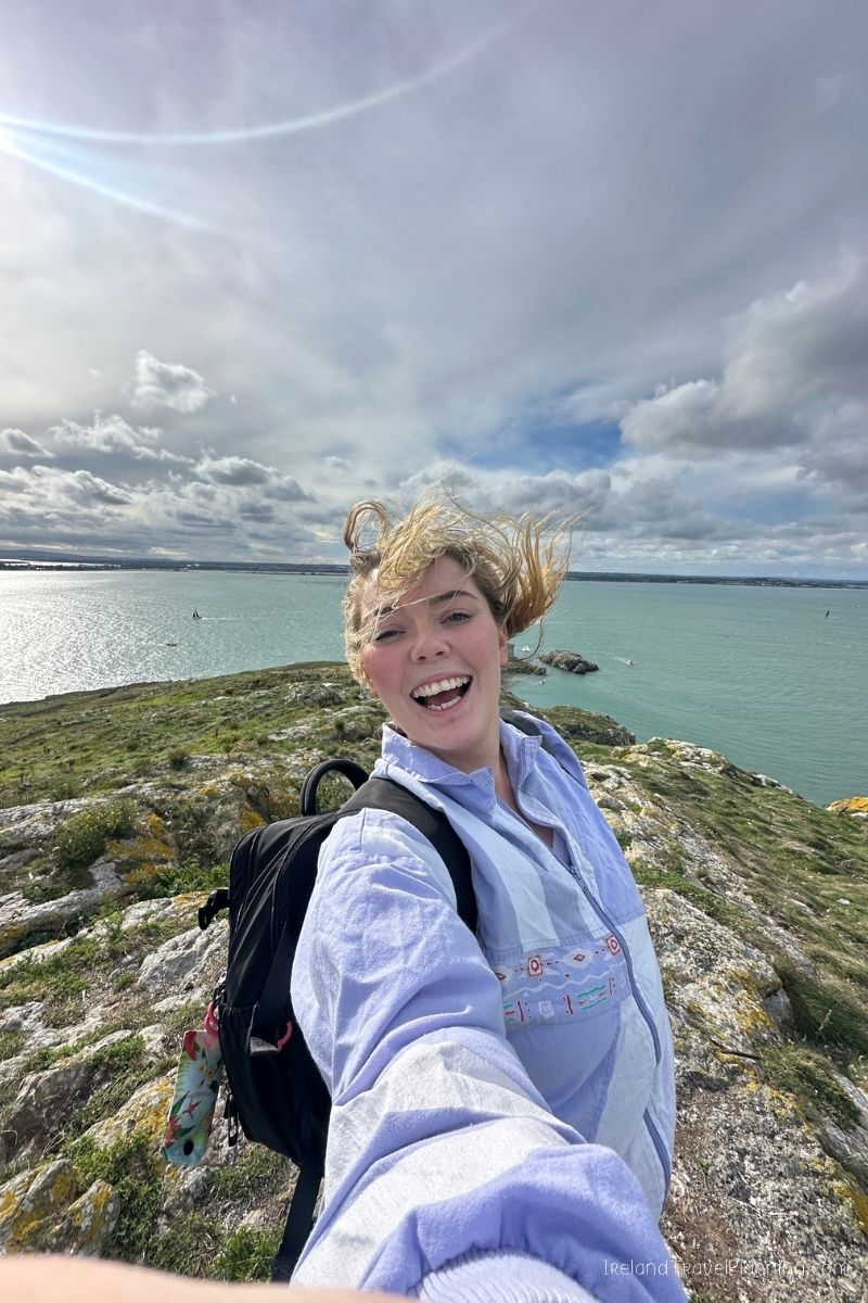 Woman with windblown hair smiling on a grassy cliff overlooking the ocean