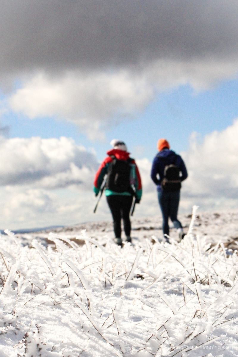 Two hikers with backpacks walk through a snowy, frosted landscape under a cloudy sky.