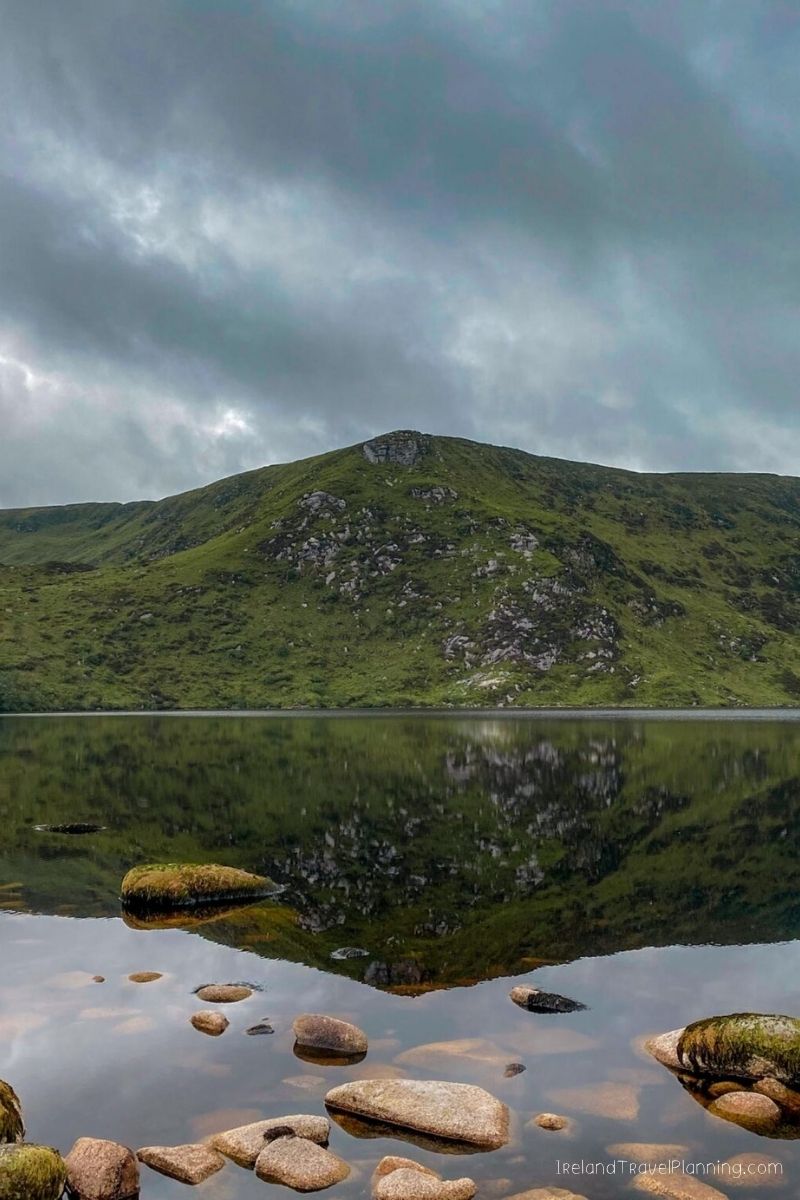 Serene Irish lake with a green mountain reflecting in calm water under a cloudy sky.
