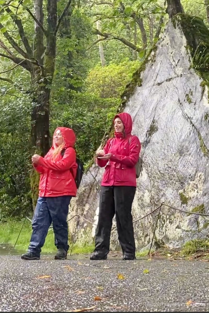 Two women in red raincoats stand near a large rock face in a forest.