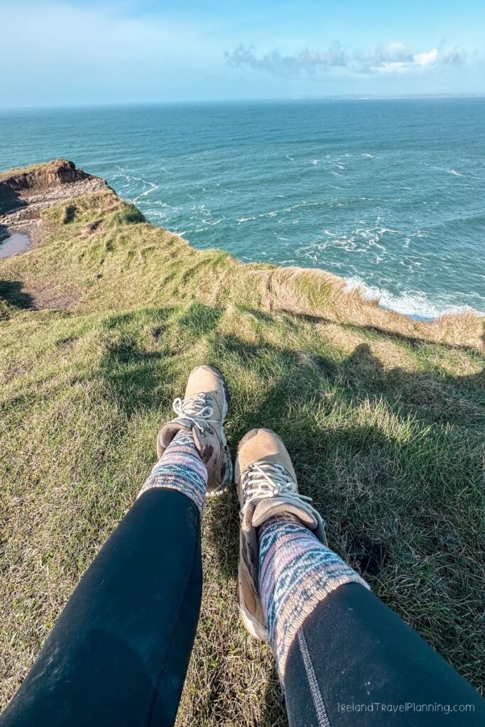 Person's legs in hiking boots and colorful socks sitting on a grassy cliff overlooking the ocean.