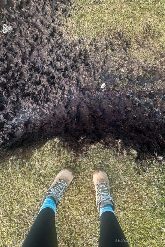 Hiking boots stand on grass next to a muddy ditch.