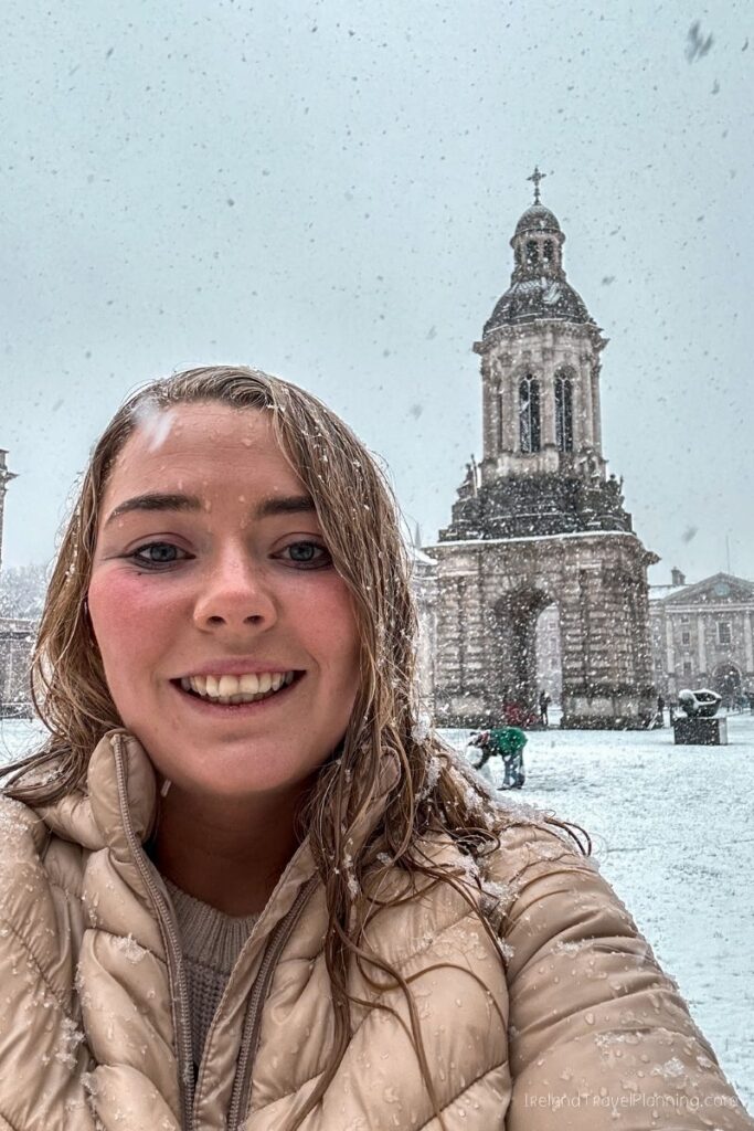 Young woman smiling in a snowy Dublin courtyard with Trinity College's campanile.