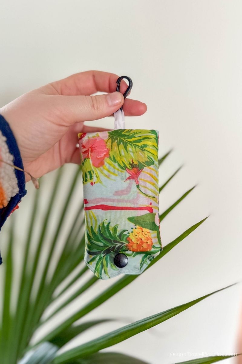 Hand holds a folded reusable shopping bag with a tropical print and carabiner clip.