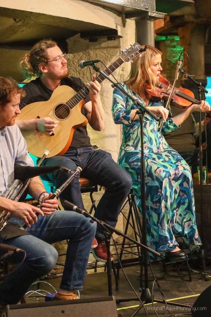 Musicians play guitar, fiddle, and bagpipes at a lively Irish pub.