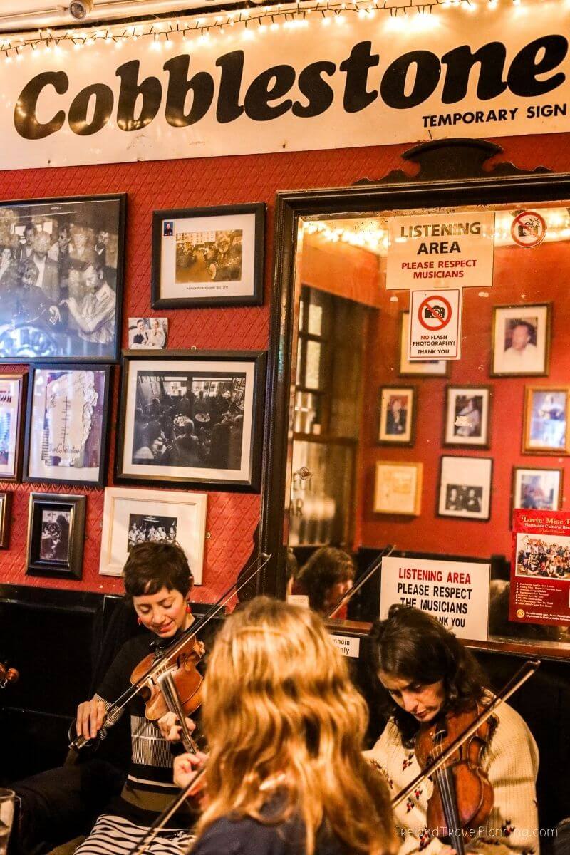 Musicians play violins at Cobblestone, a venue with framed photos and a "Listening Area" sign.