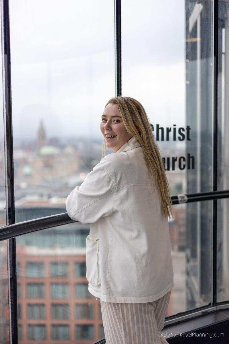 Woman in white jacket smiles at camera with city view behind her.