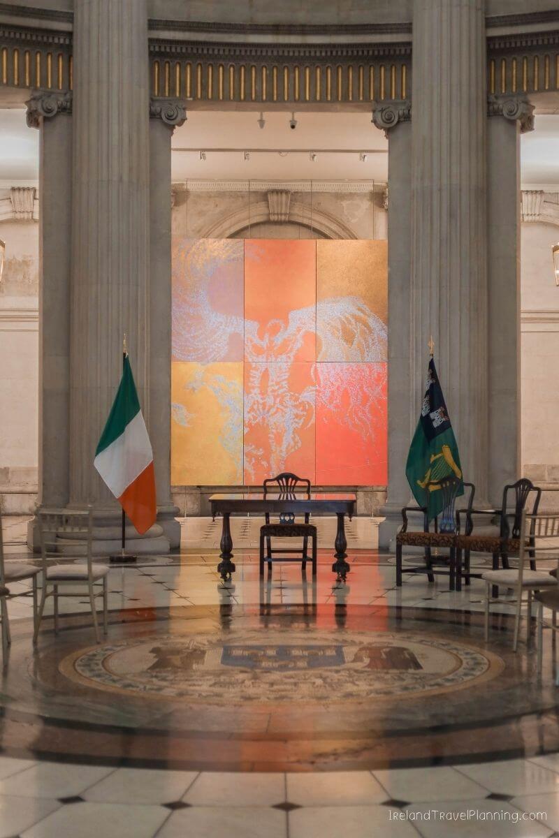 Irish flags flank a table and chairs before a large abstract artwork in a grand hall.